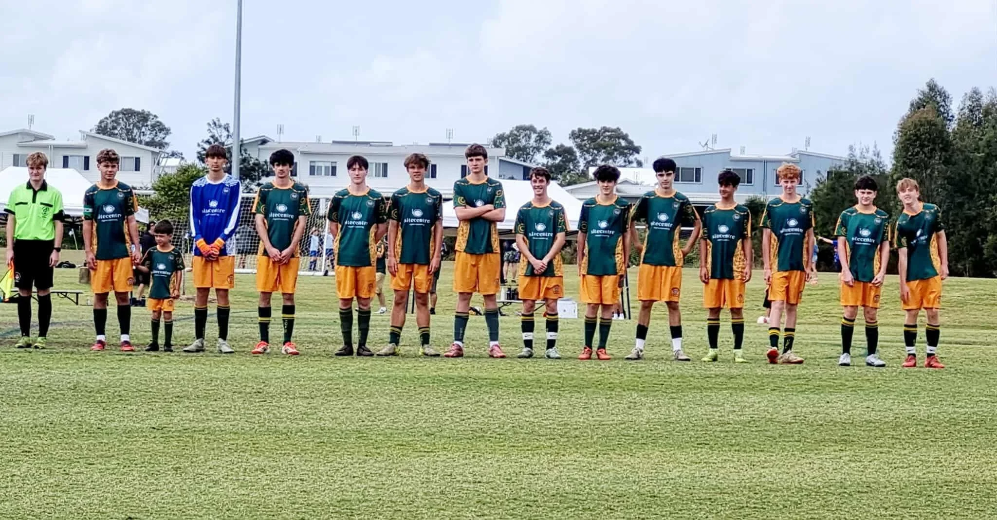 A youth soccer team standing in a line on a field with two goalposts in the background, wearing green and yellow jerseys and black shorts.