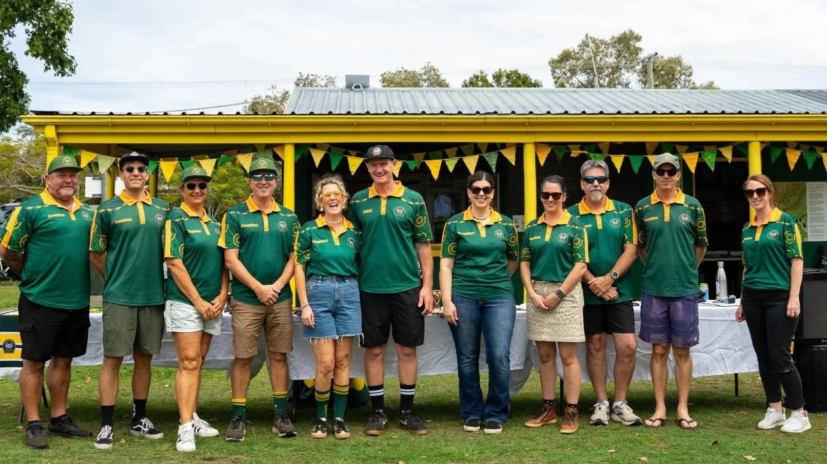 A group of eleven people standing outdoors in front of a yellow building decorated with green and yellow bunting. They are wearing matching green and yellow sports shirts, some with hats and sunglasses, and are smiling.