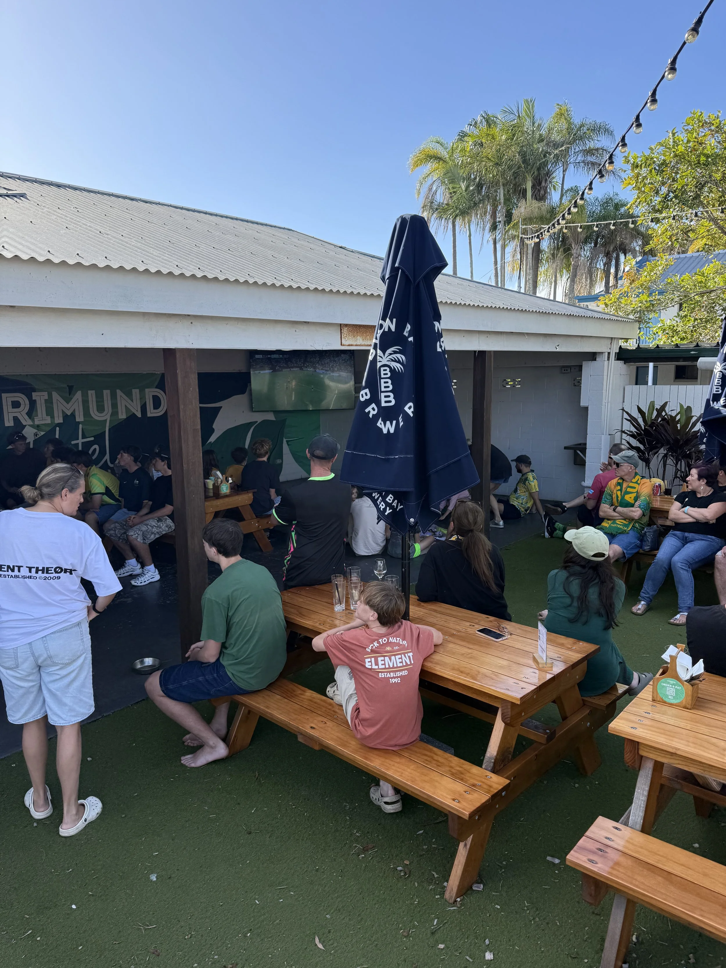 People gathered outside at a food venue with a white building, tall palm trees, string lights, and wooden picnic tables on artificial grass.