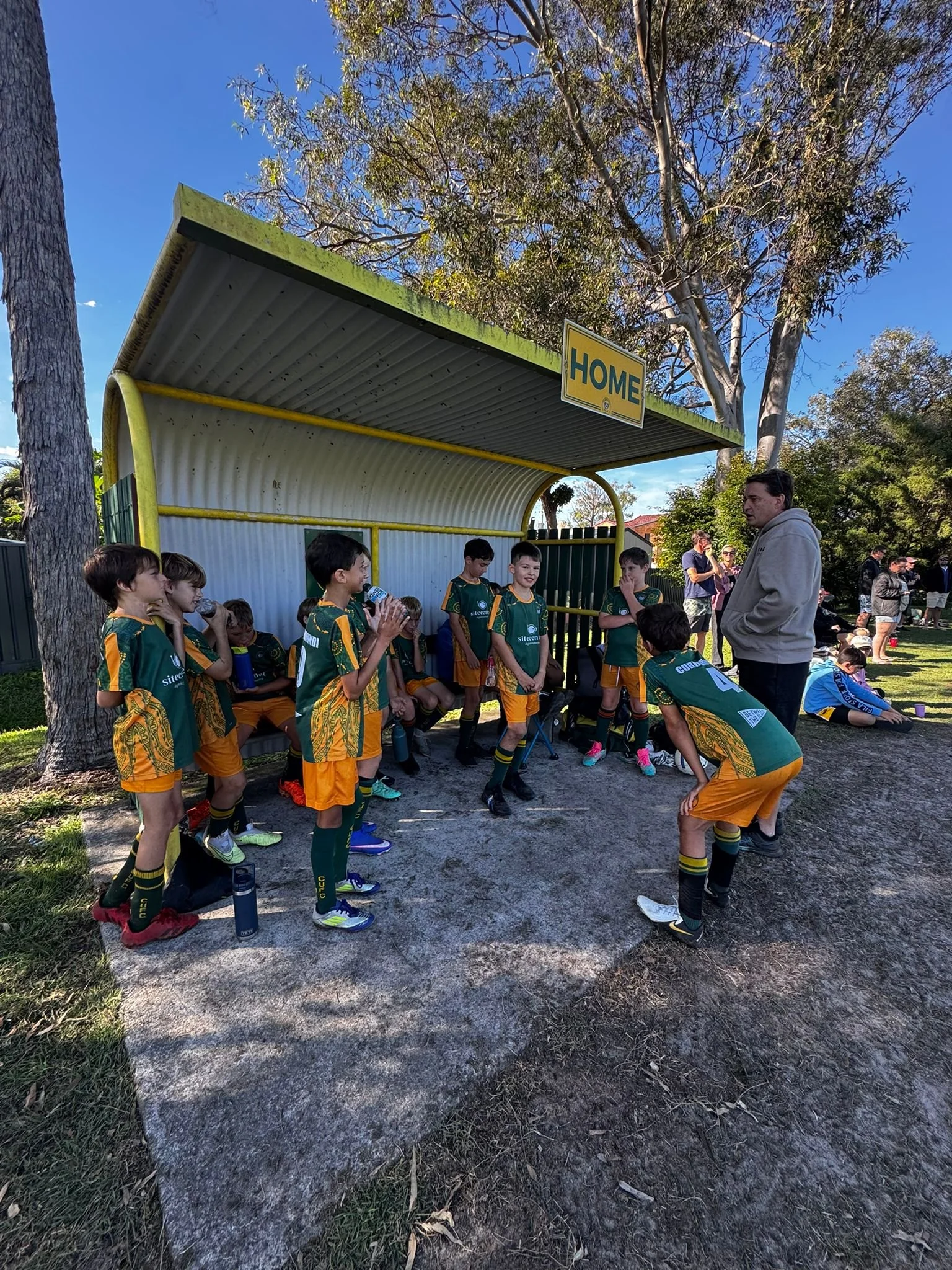A group of children in green and gold sports uniforms sitting and standing under a bus stop shelter labeled 'HOME' in a park, with a man standing in front of them, talking. There are trees and a few adults in the background.