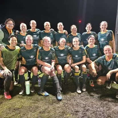 Team of women soccer players in green and black uniforms, posing for a group photo after a match, on a dark outdoor field.
