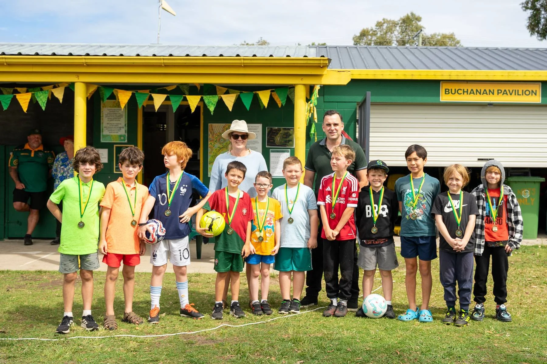 A group of children with medals around their necks poses outside a building labeled 'Buchanan Pavilion,' with two adults standing behind them, holding soccer balls, some wearing sports jerseys, and outdoor decorations in the background.