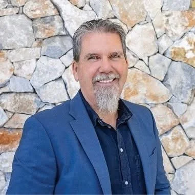 A man with gray hair and a beard smiling, wearing a blue blazer and a dark shirt, standing in front of a stone wall.