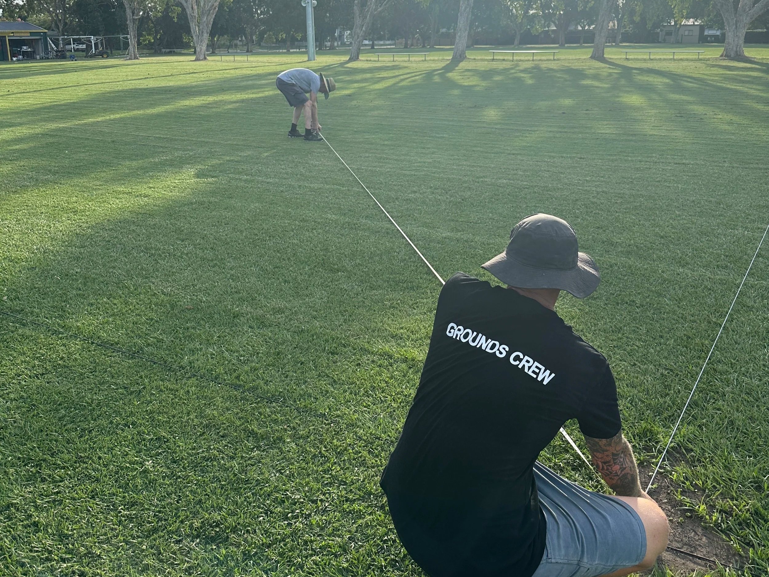 A person in a black shirt with white text on the back, gray shorts, and a large floppy hat is sitting on the grass holding a string or cord. In the background, another person in a hat, gray shirt, and shorts is walking or bending down on the grassy field, possibly playing golf or engaging in an outdoor activity. The scene is in a park with trees and benches.