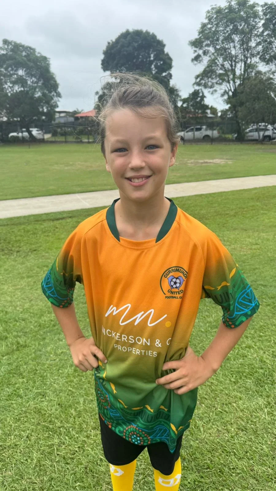 A young girl in a colorful sports uniform standing on a grassy field with hands on hips, smiling at the camera.