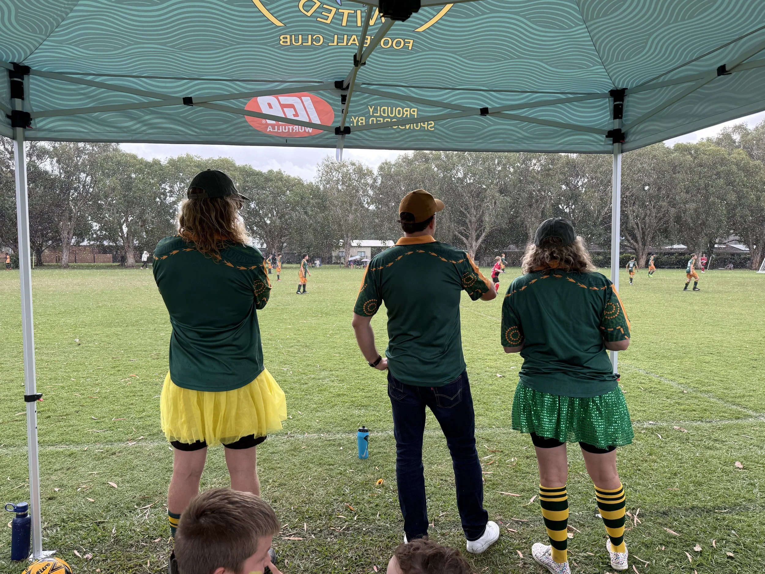 Three people standing under a canopy watching a rugby game on a grass field. Two women and one man, dressed in colorful sports attire, with trees and more players visible in the background.