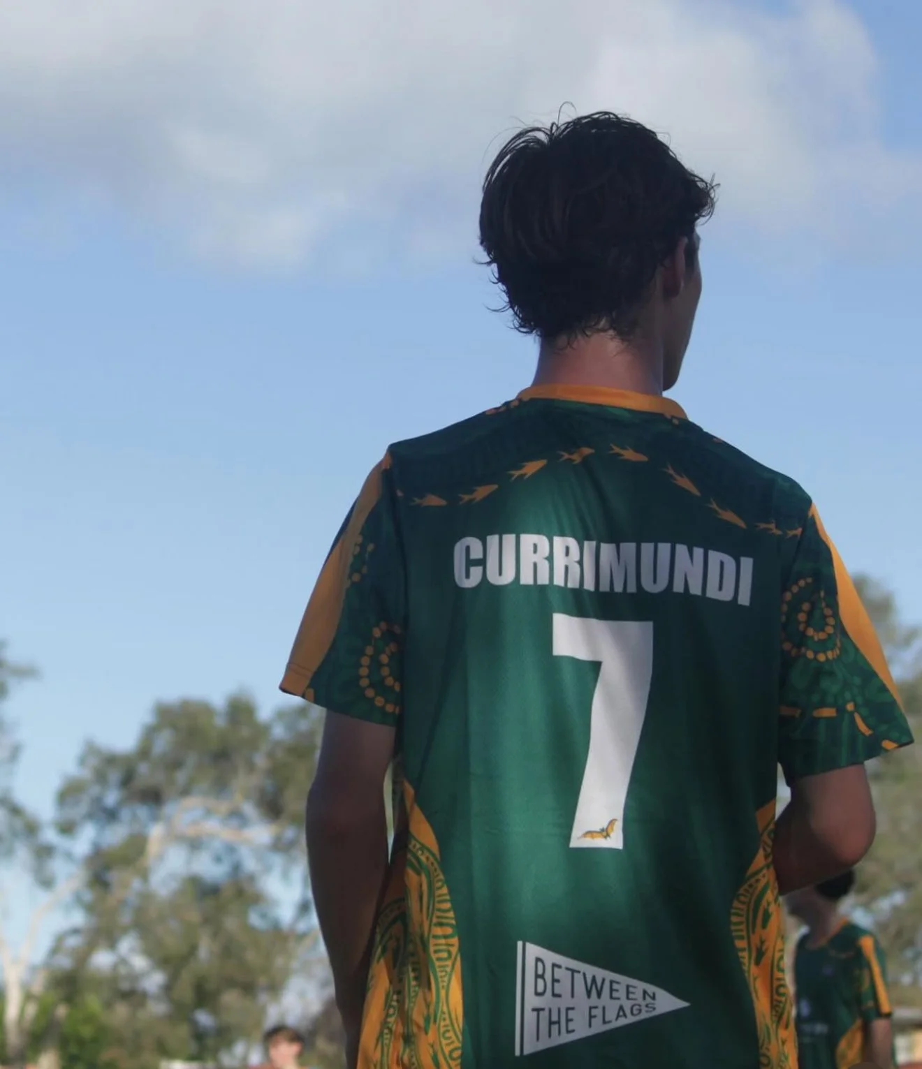 A person wearing a green and yellow sports jersey with the name 'Currimundi' and the number 7 seen from the back, standing outdoors with trees and a blue sky in the background.