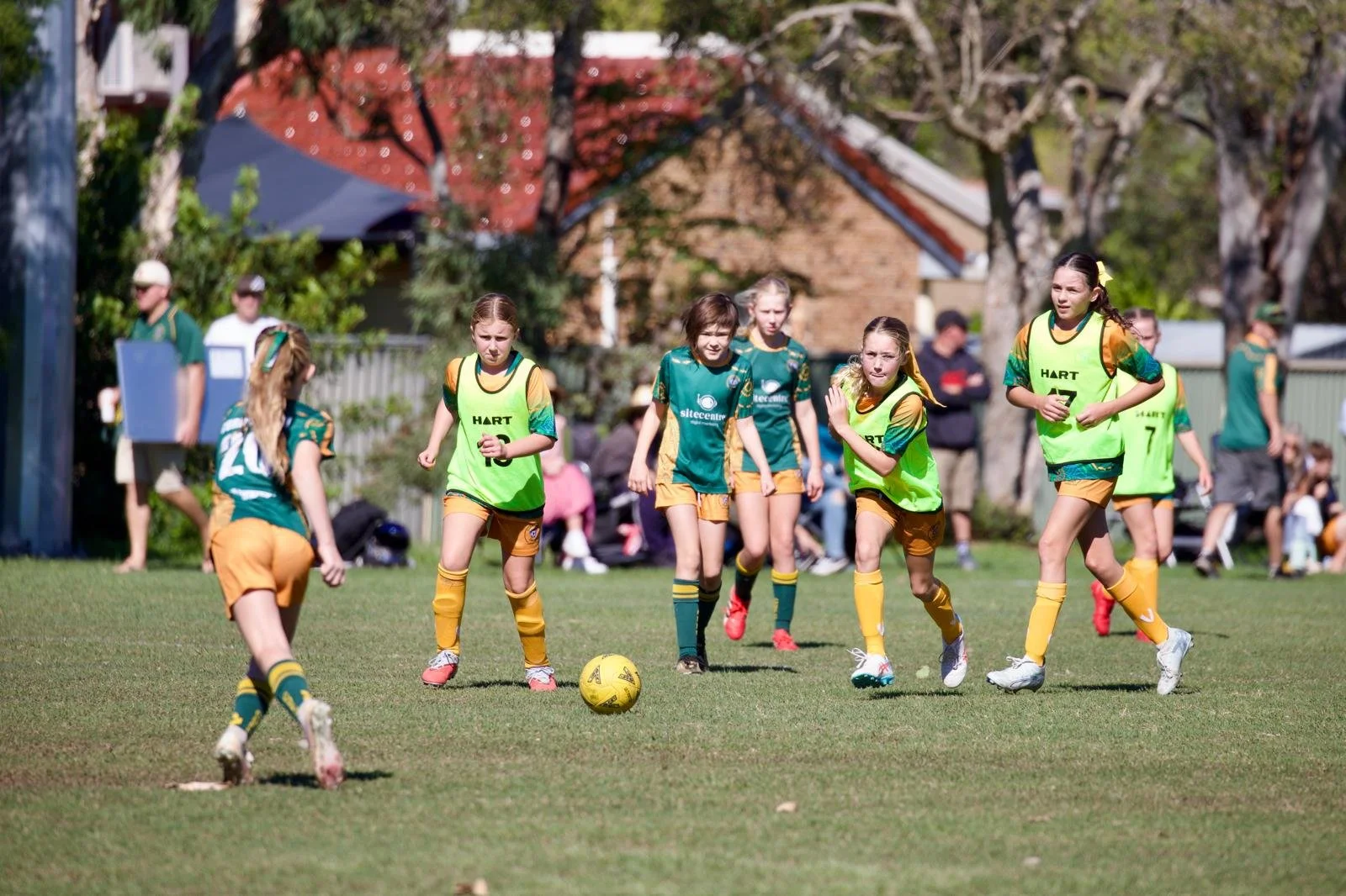 Girls playing soccer on a grassy field, wearing green and yellow uniforms, with one girl kicking a yellow soccer ball.