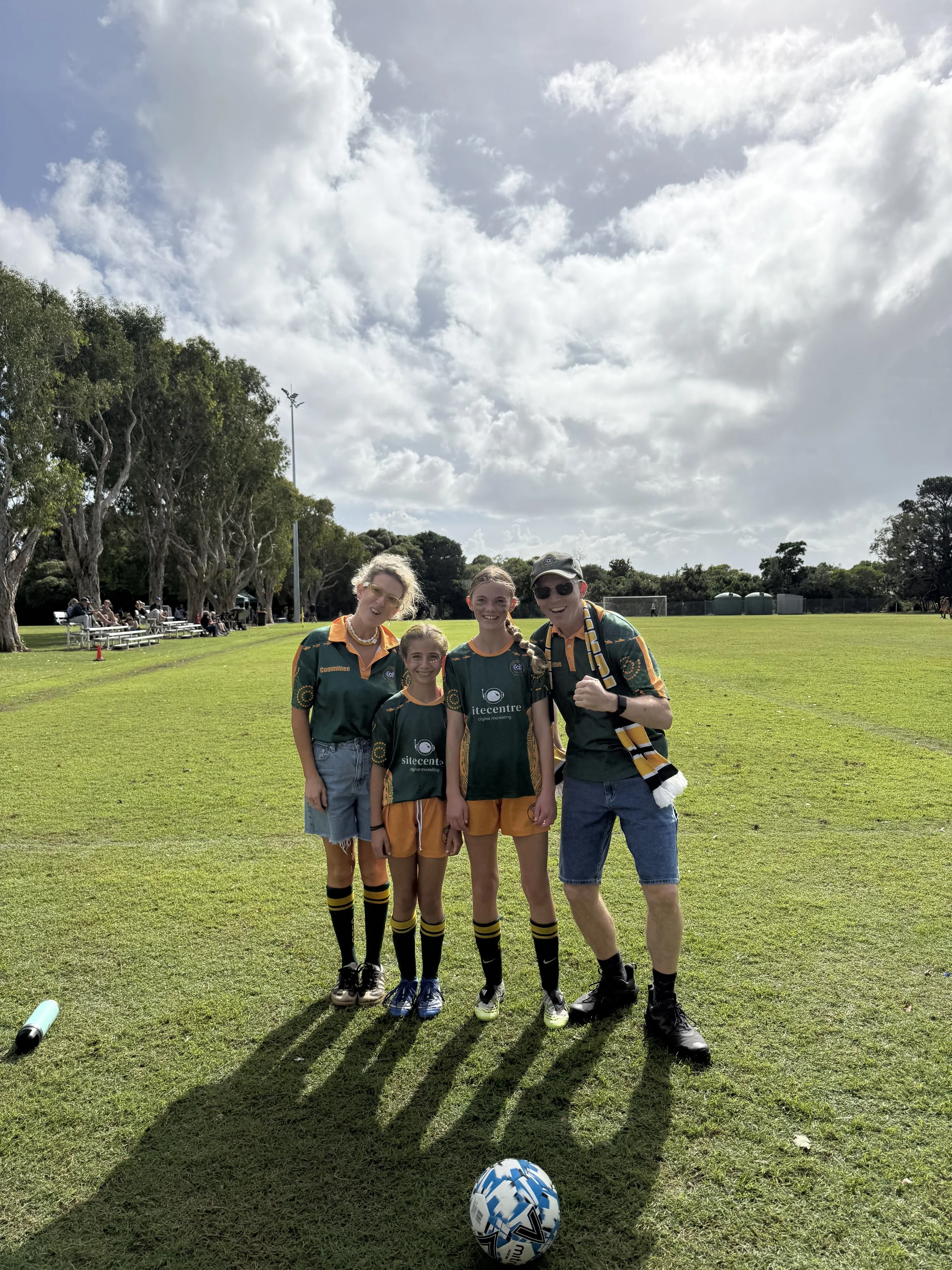 Four people in sports attire standing on a grassy field with trees and benches in the background. Two are young girls, and two are adults. All are smiling and posing for a photo, with a soccer ball on the ground in front of them.