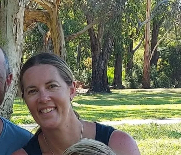 A smiling woman with light brown hair, wearing hoop earrings and a black sleeveless top, outdoors with trees and grass in the background.