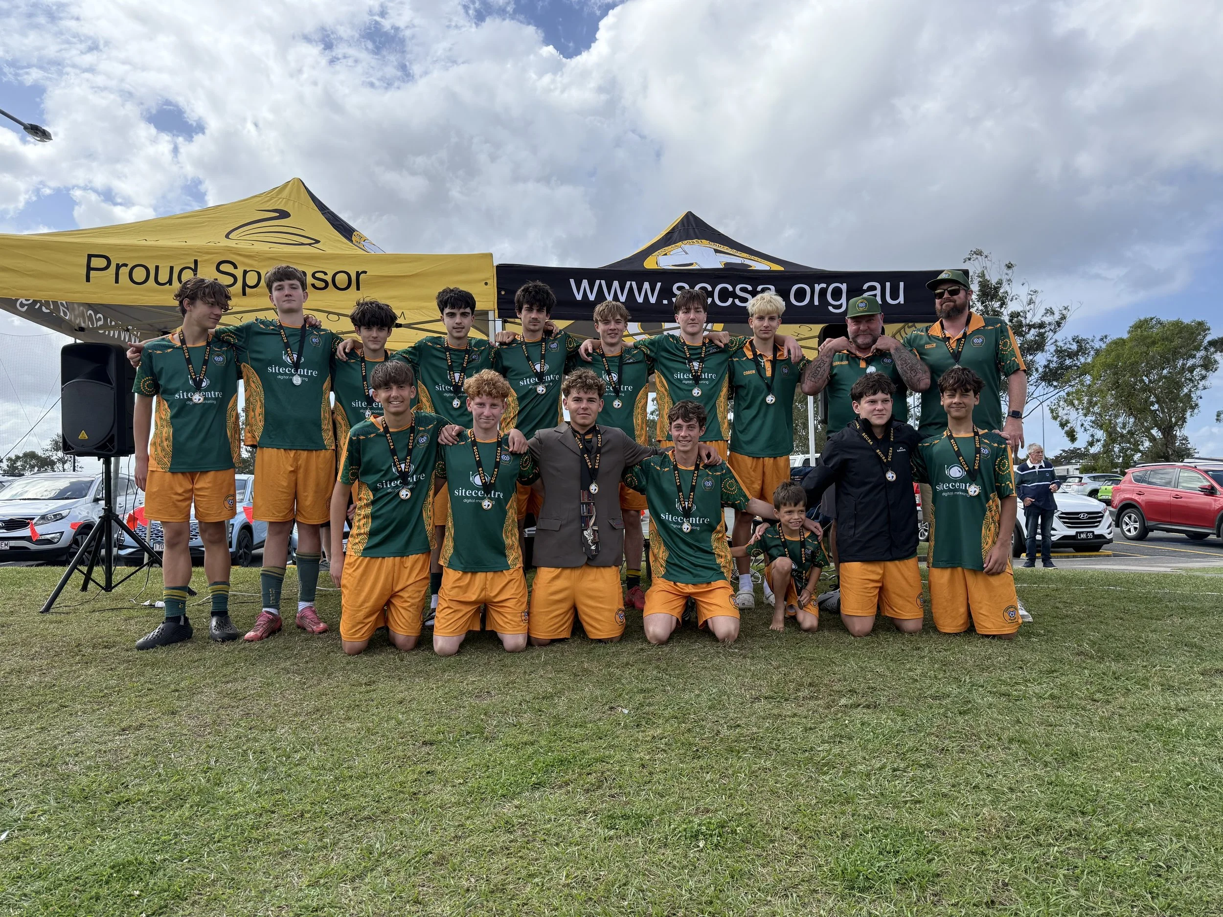 A group of young male athletes and their coaches wearing green and yellow sports jerseys and shorts, standing on a grassy field under a partly cloudy sky, posing for a victory photo with medals around their necks. They are in front of a tent with banners for sponsors.