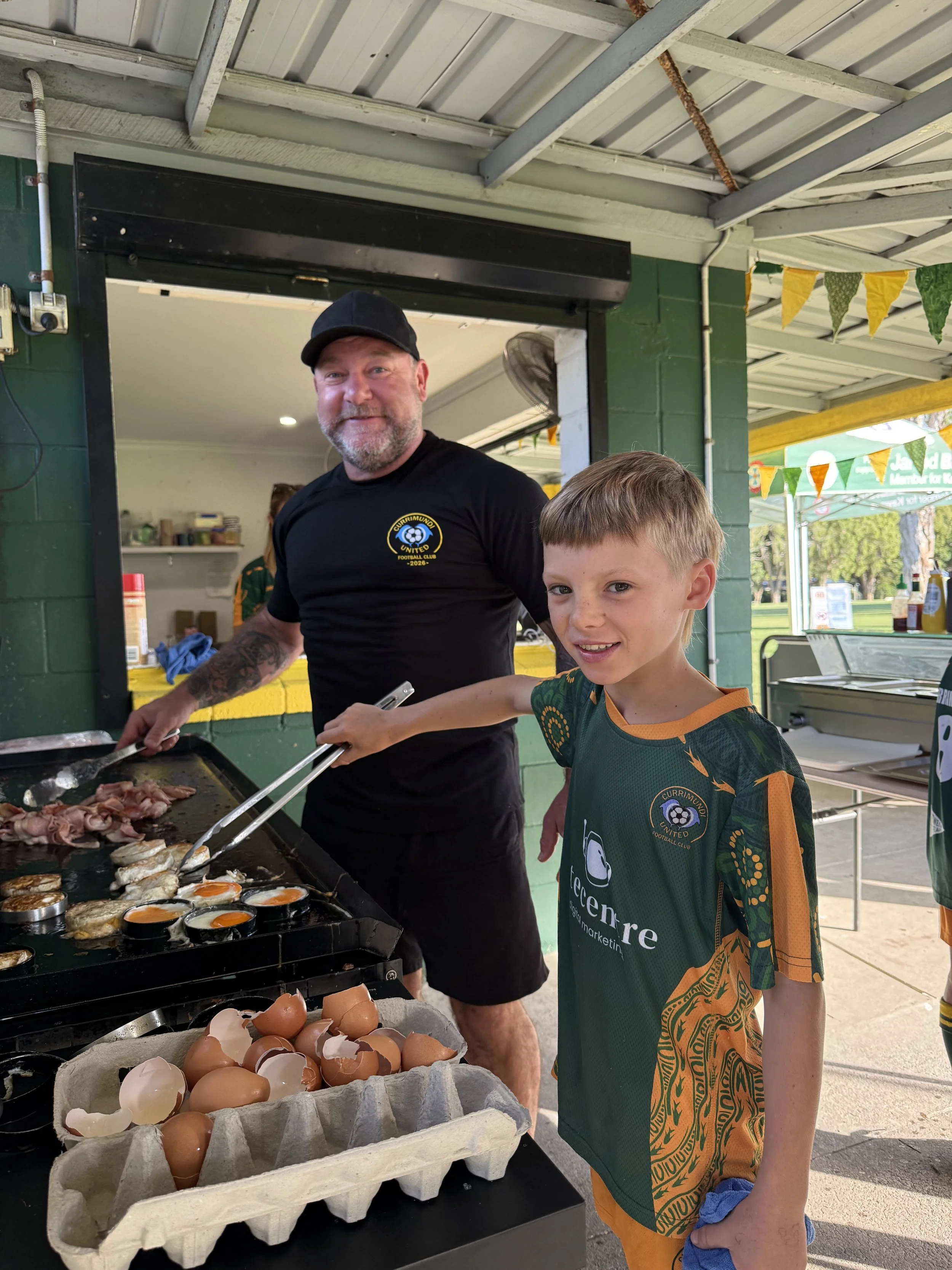 A man and a boy are cooking breakfast outdoors at a market or fair. The man is cooking eggs and bacon on a griddle, while the boy is holding a pair of tongs. There are eggshells in a carton in front of them, and the area is decorated with yellow and green bunting.