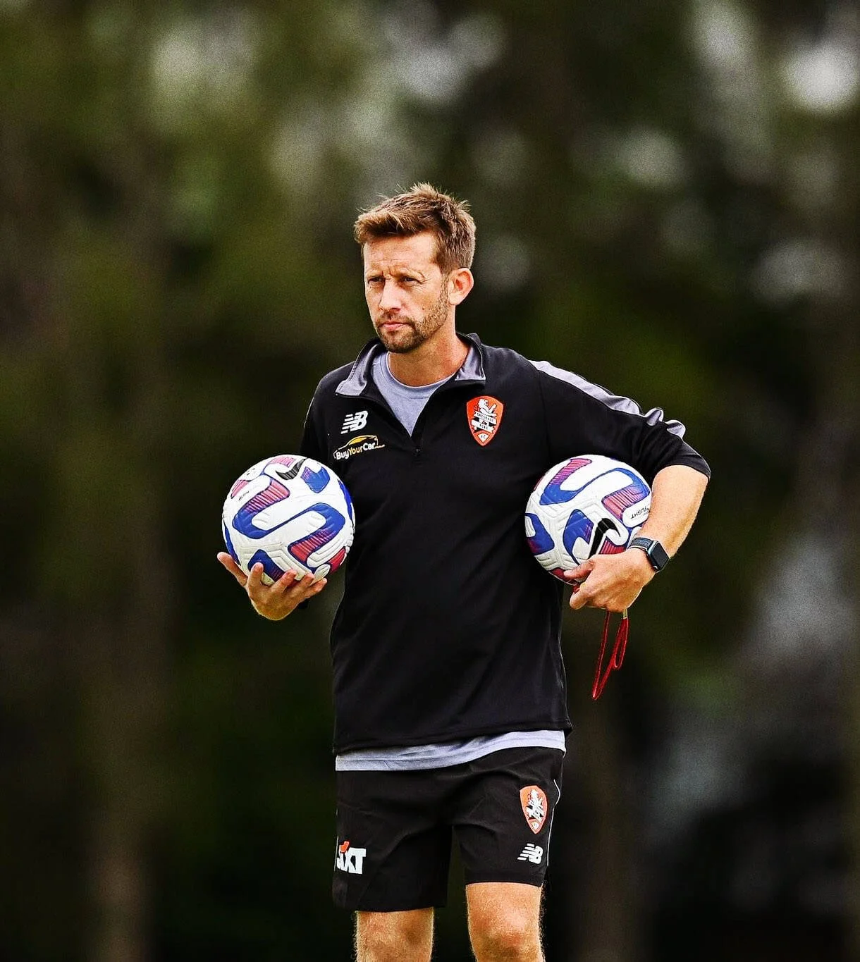 A man in black sportswear holding two soccer balls during a practice or game outdoors.