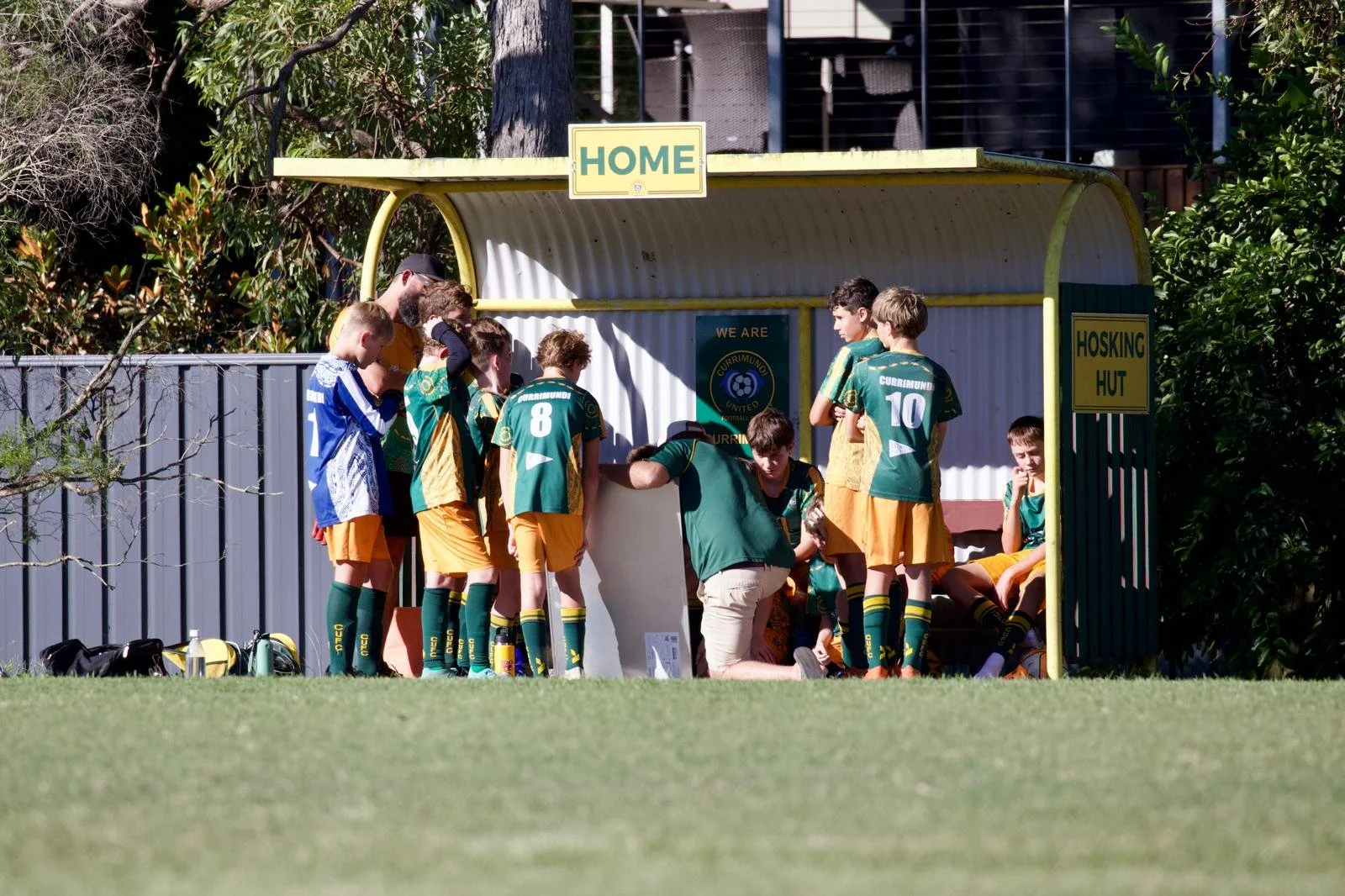 Youth soccer team huddled under a shelter on a soccer field, with a coach giving instructions. The players wear green and yellow uniforms, with some seated and others standing. Sports bags and water bottles are on the ground.