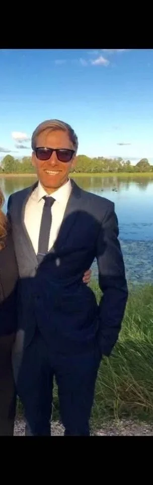 A man wearing a suit and sunglasses standing outdoors near a body of water with trees in the background.