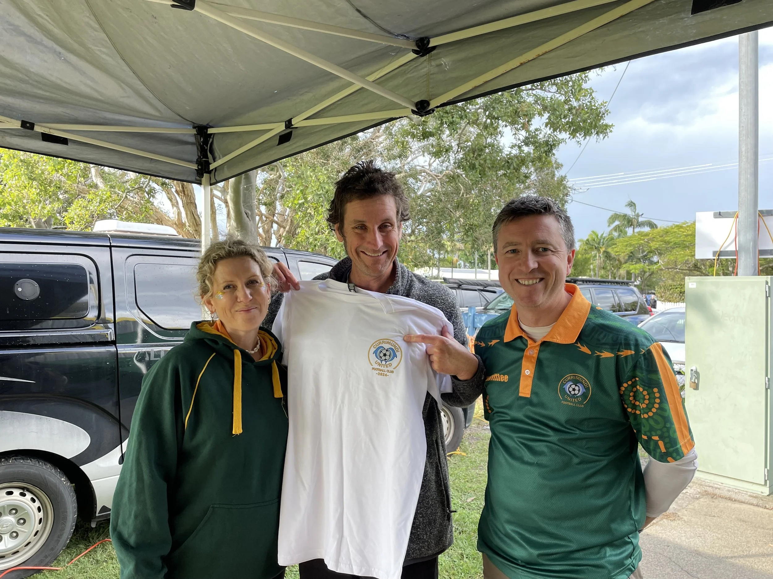 Three people standing outdoors under a canopy, smiling and holding up a sports jersey with a logo that says 'Currimundoo United Football Club 2022'.
