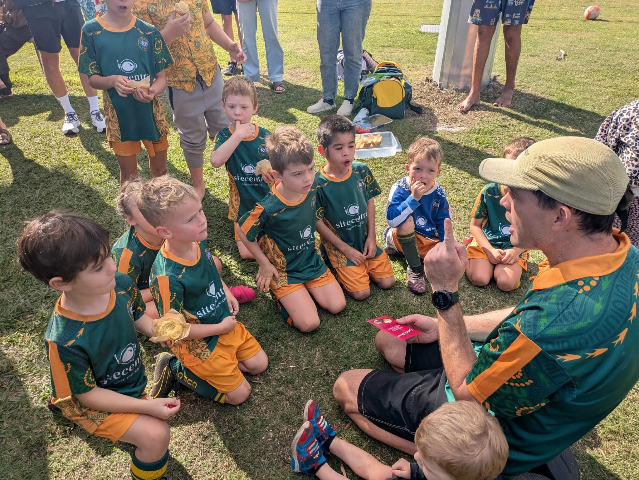 Group of children and adults gathered outdoors on grass, some children are sitting while others are standing; children are wearing sports uniforms with a logo, some hold bread or snacks, and adults are engaging with them.