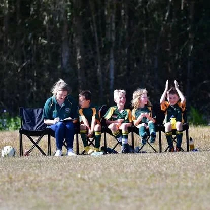 Four children in soccer uniforms sitting on chairs on a grassy field. A woman sits on a chair to their left. A soccer ball is on the ground nearby. Trees are in the background.