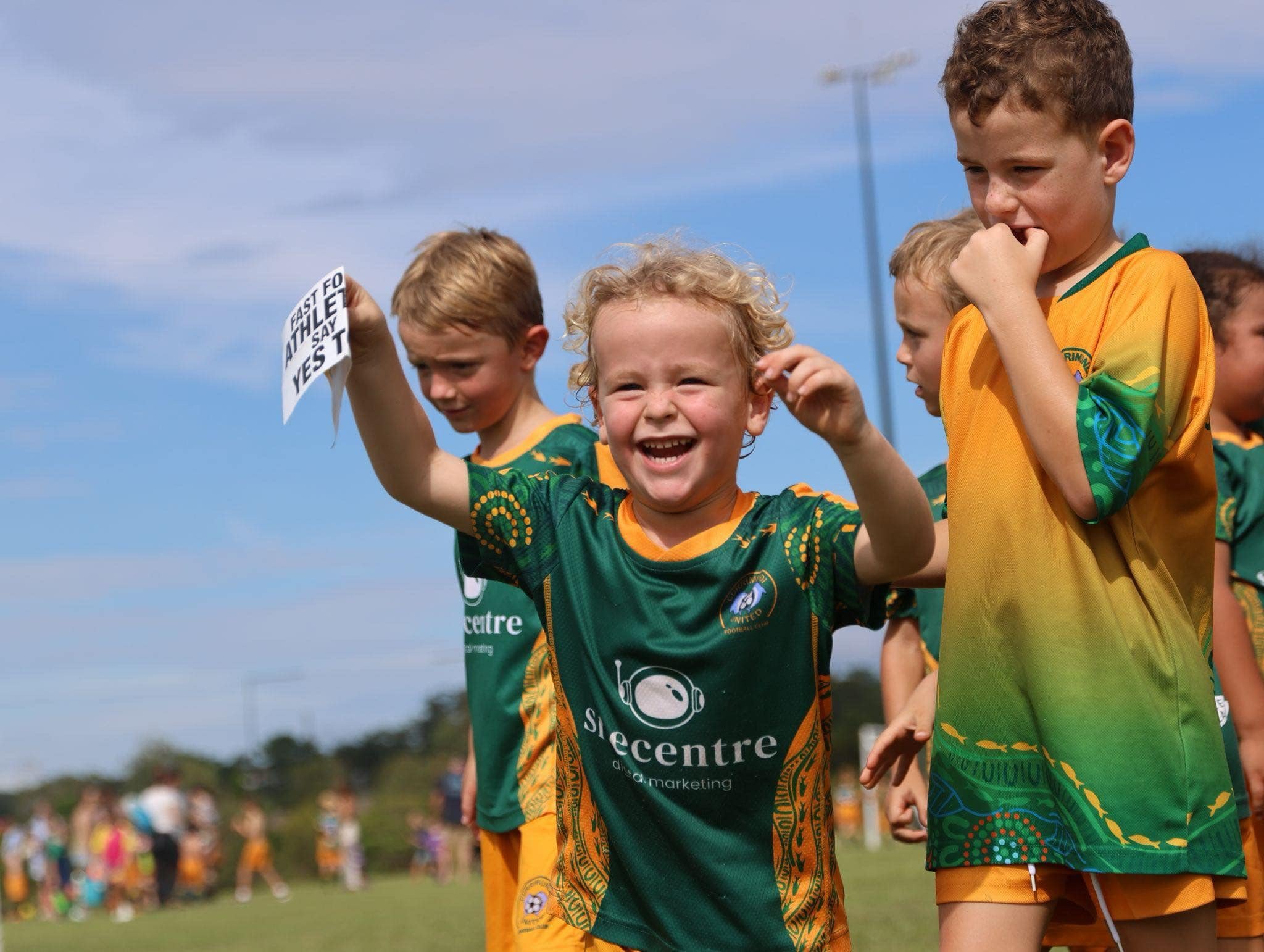 Group of children in sports jerseys standing outdoors at a sporting event, with one child holding a sign and others adjusting their hat or hair, under a partly cloudy sky.