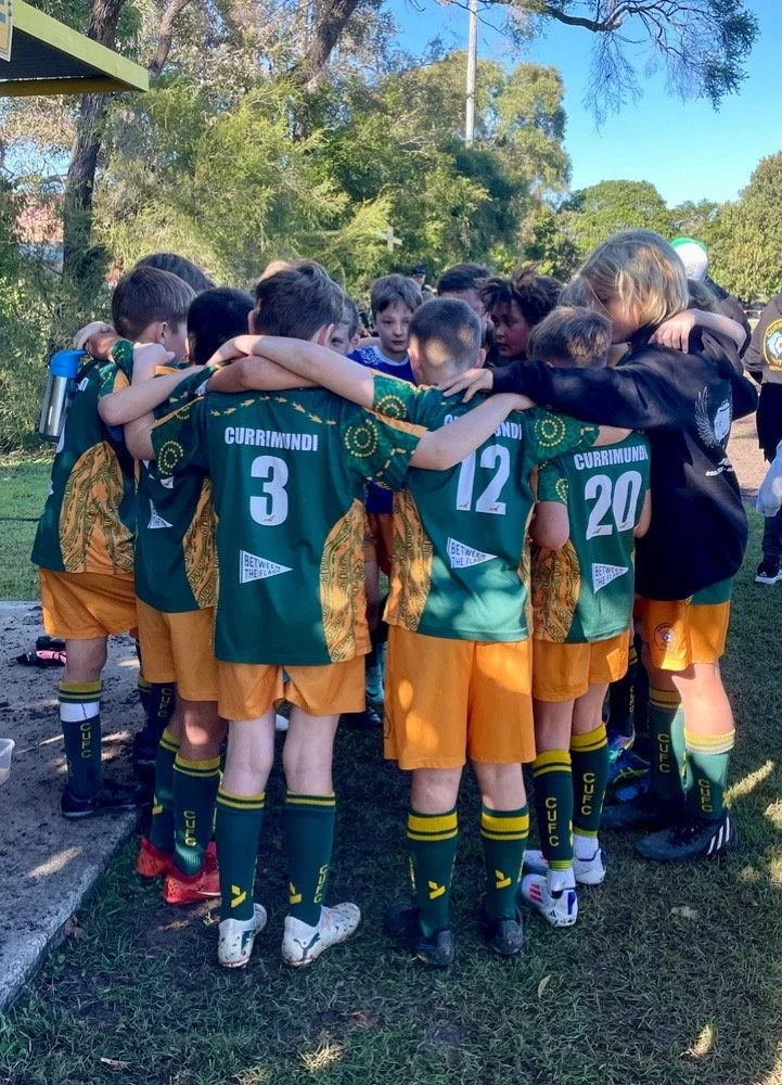 Youth rugby team standing in a tight circle, huddled with arms around each other, outdoors on grass with trees in the background.