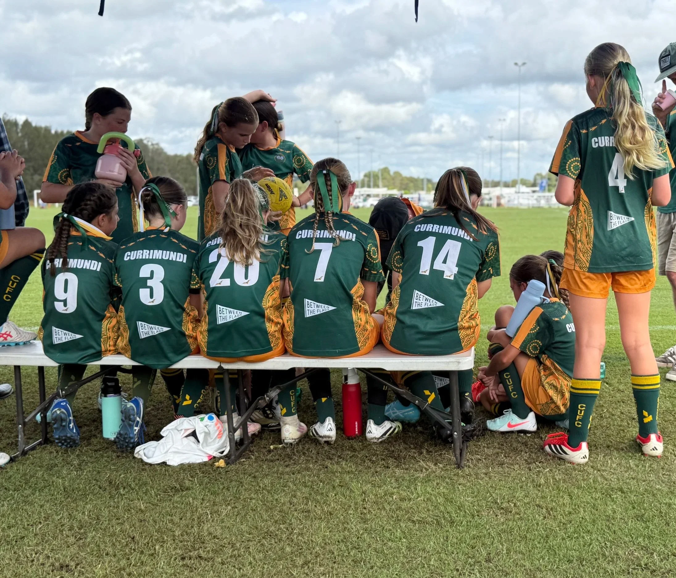 Young female athletes sitting and standing on a bench during a sports event, wearing green and yellow uniforms with numbers on the back, on a grassy field with a cloudy sky in the background.