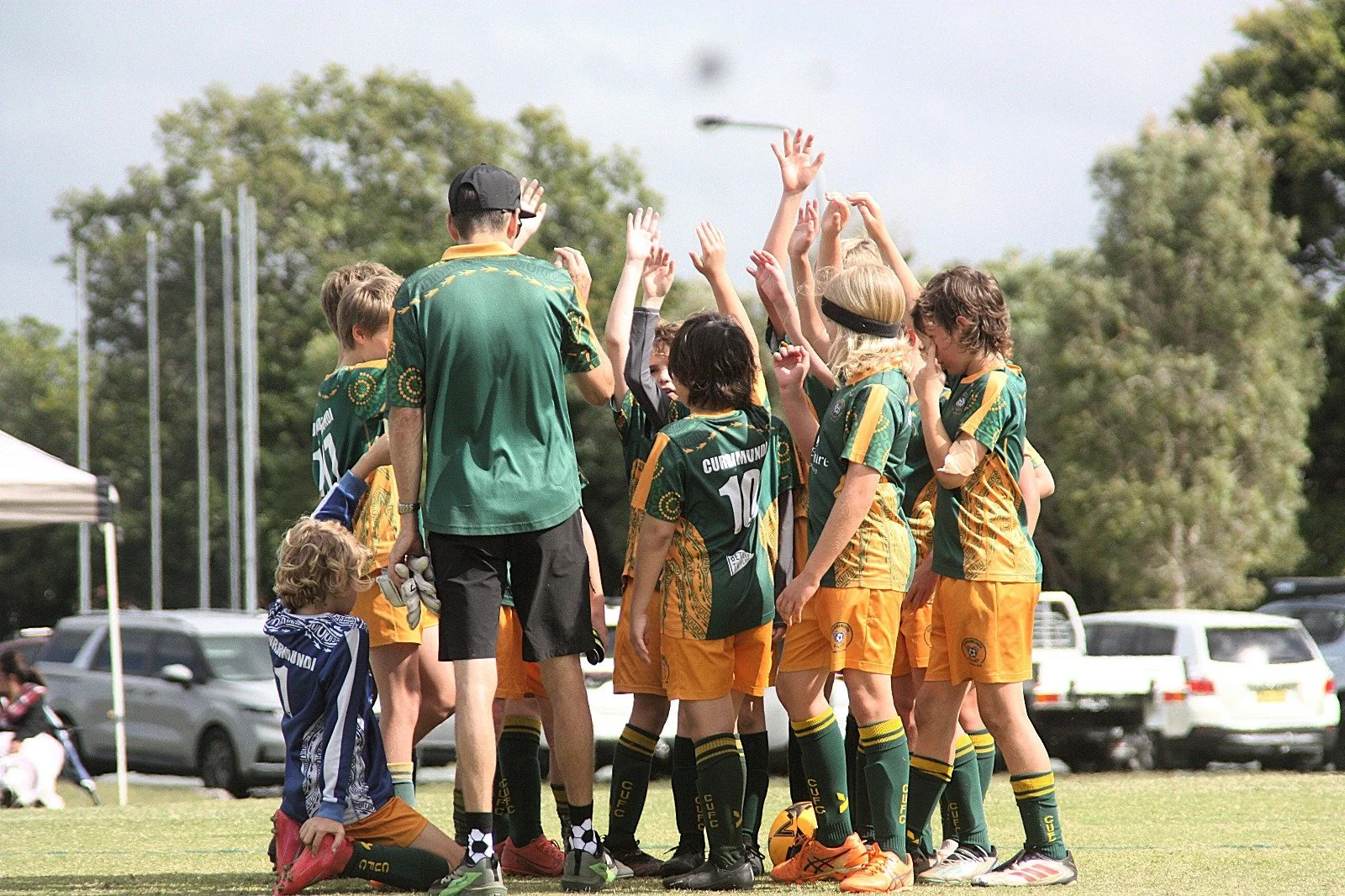 A group of young children and a coach on a soccer field, giving each other high-fives after a game or practice. The children are wearing green and yellow soccer uniforms.
