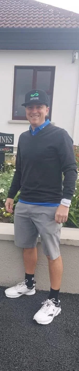 A man standing outdoors in front of a house with a window and garden, smiling, wearing a baseball cap, a black sweater over a blue shirt, gray shorts, black socks, and white sneakers.