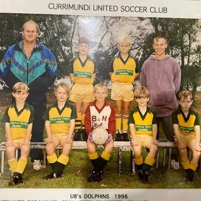 Group photo of the Currimundi United Soccer Club U8's Dolphins team from 1996. The team includes seven young girls wearing yellow and green soccer uniforms and a girl in a red and white jersey holding a soccer ball. Two adults, likely coaches or trainers, stand behind the team, one on each side. The picture is taken outdoors on a grassy field with trees in the background.