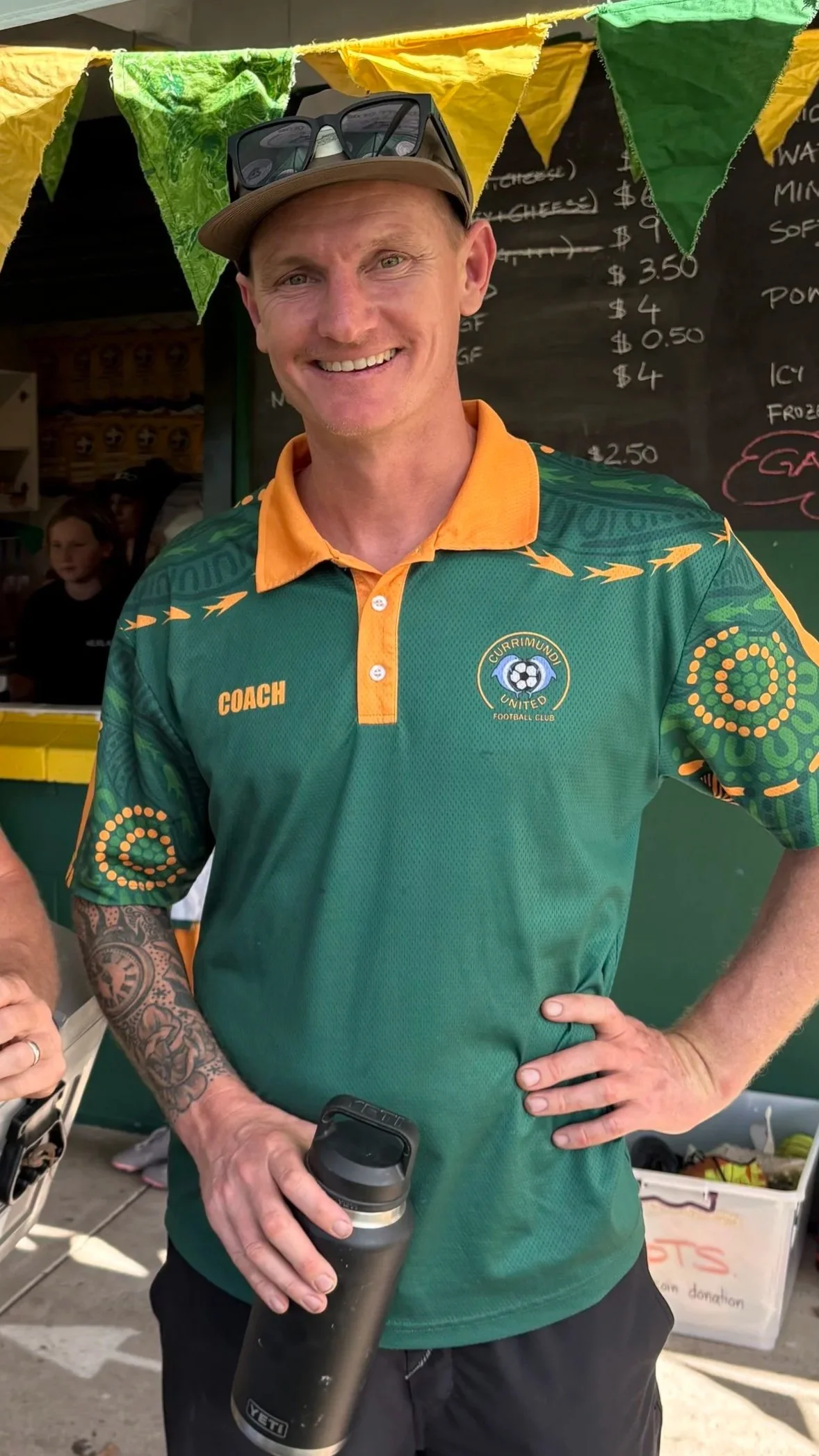 Man smiling, wearing a green and orange sports shirt with the logo of Currimundi United Football Club, holding a black drink tumbler, standing outdoors at a food stall with yellow and green bunting overhead and a chalkboard menu behind him.