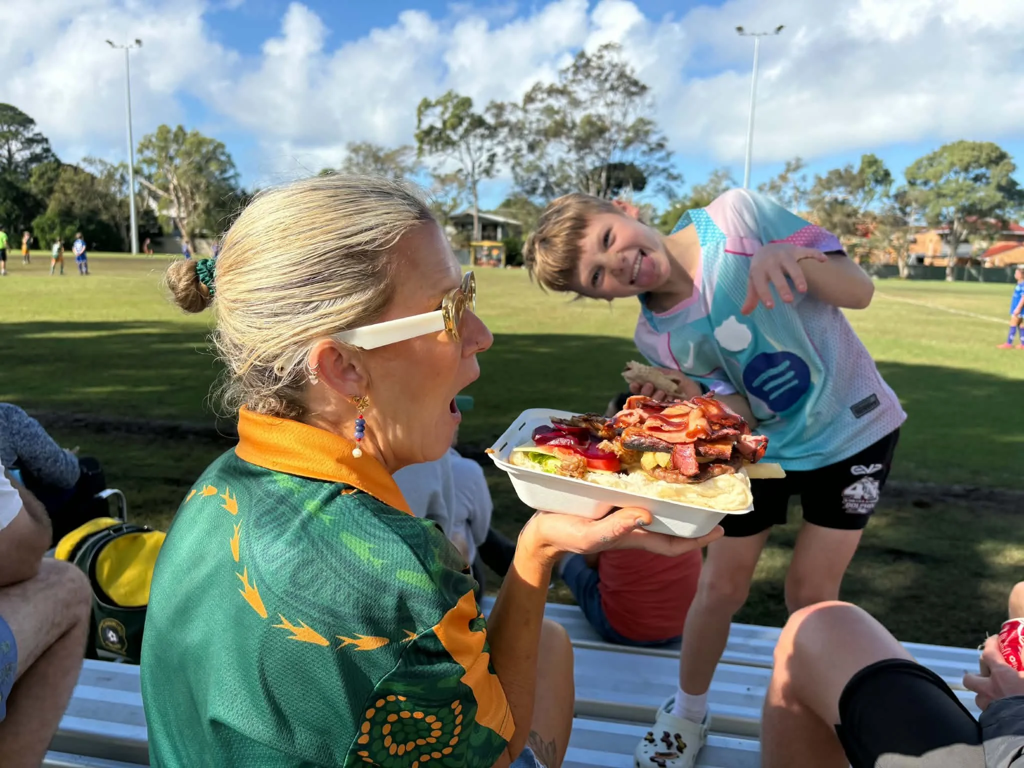 A woman with blonde hair in a bun, wearing glasses and a colorful shirt, holding a tray of food, with a girl in a sports jersey smiling and pointing at her, in an outdoor park with children playing and trees under a blue sky.