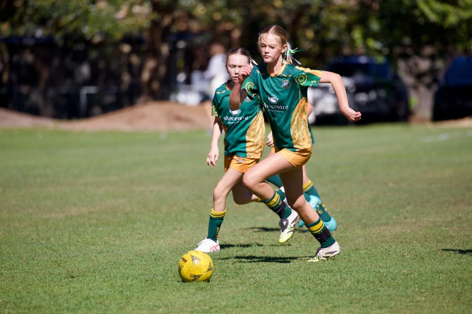 Two young girls in green and yellow soccer uniforms playing soccer on a grassy field, with one girl in front dribbling a yellow soccer ball and the other girl chasing behind.