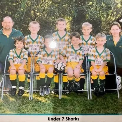 Group of young soccer players in green and yellow jerseys with two coaches, seated outdoors on chairs, with a trophy between the two middle children. The team is called 'Under 7 Sharks'.