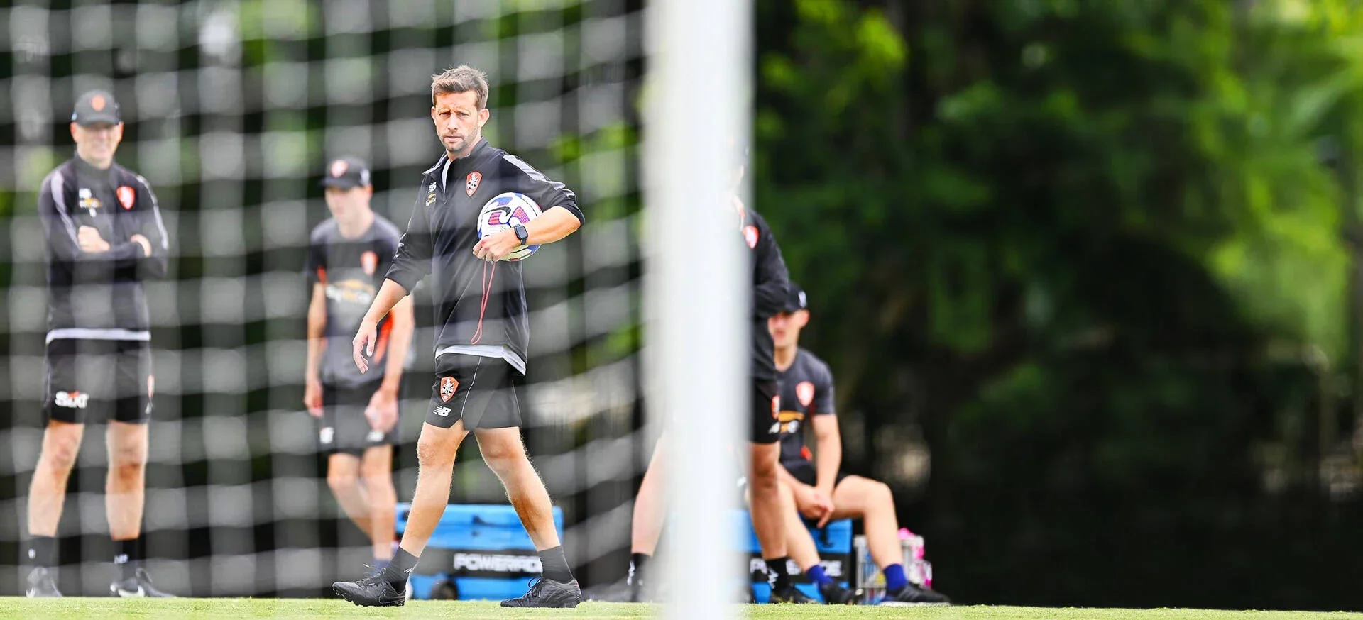 Soccer coach holding a ball on the field during practice, with players sitting on the sideline and the net in the foreground, surrounded by green trees.