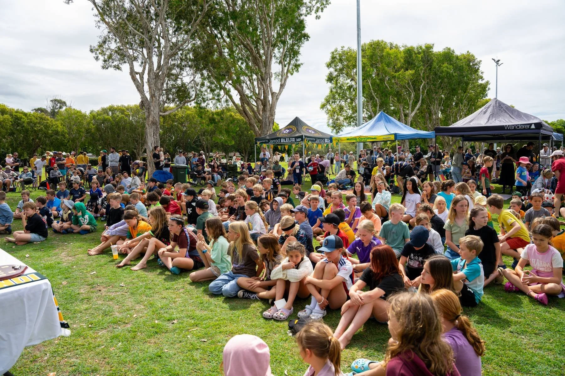 A large group of children sitting on the grass at an outdoor event, with some standing and some seated, under tall trees and tents.