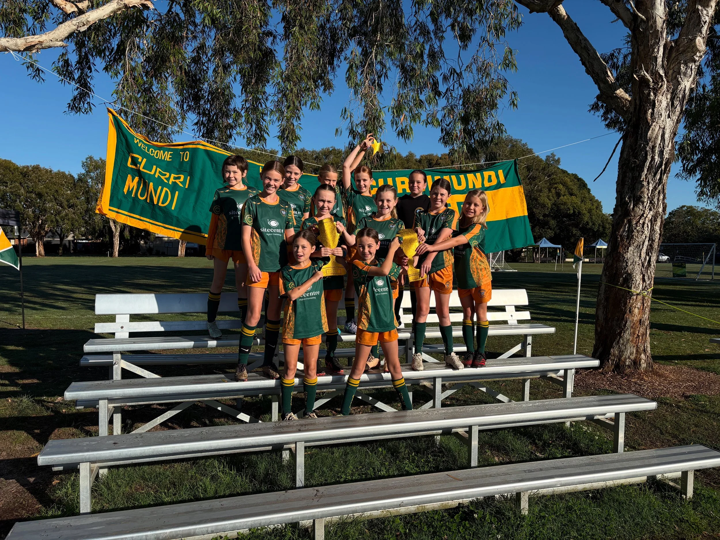 A group of young girls in green and yellow sports uniforms holding trophies, standing on bleachers outdoors, with a large tree and a green banner that reads "Welcome to Curry Mundi" in the background.