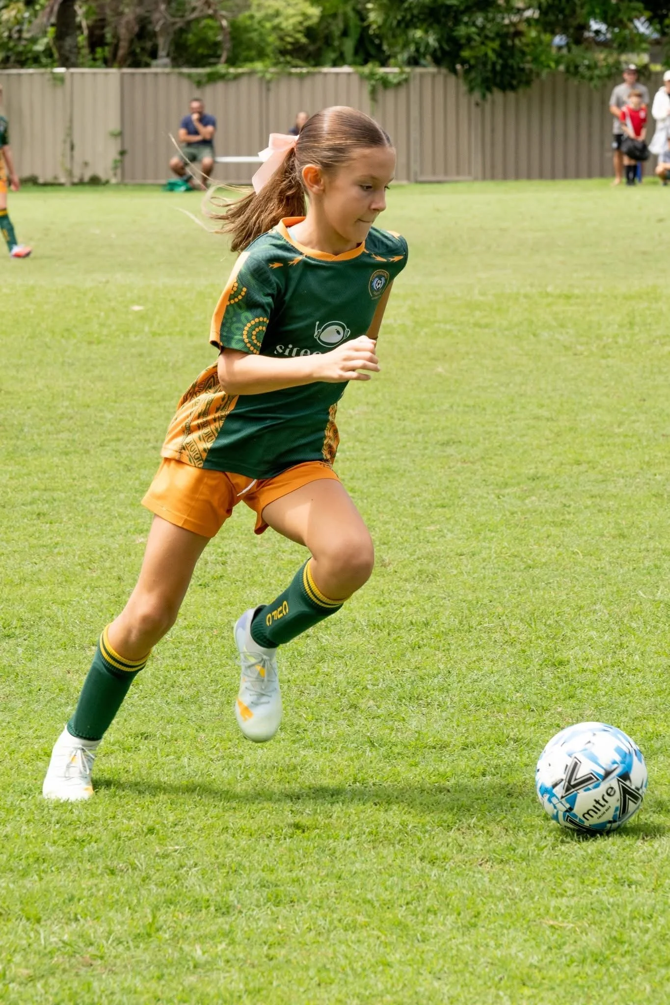 Young girl playing soccer on a grassy field, wearing a green and yellow sports uniform, and running towards a soccer ball.