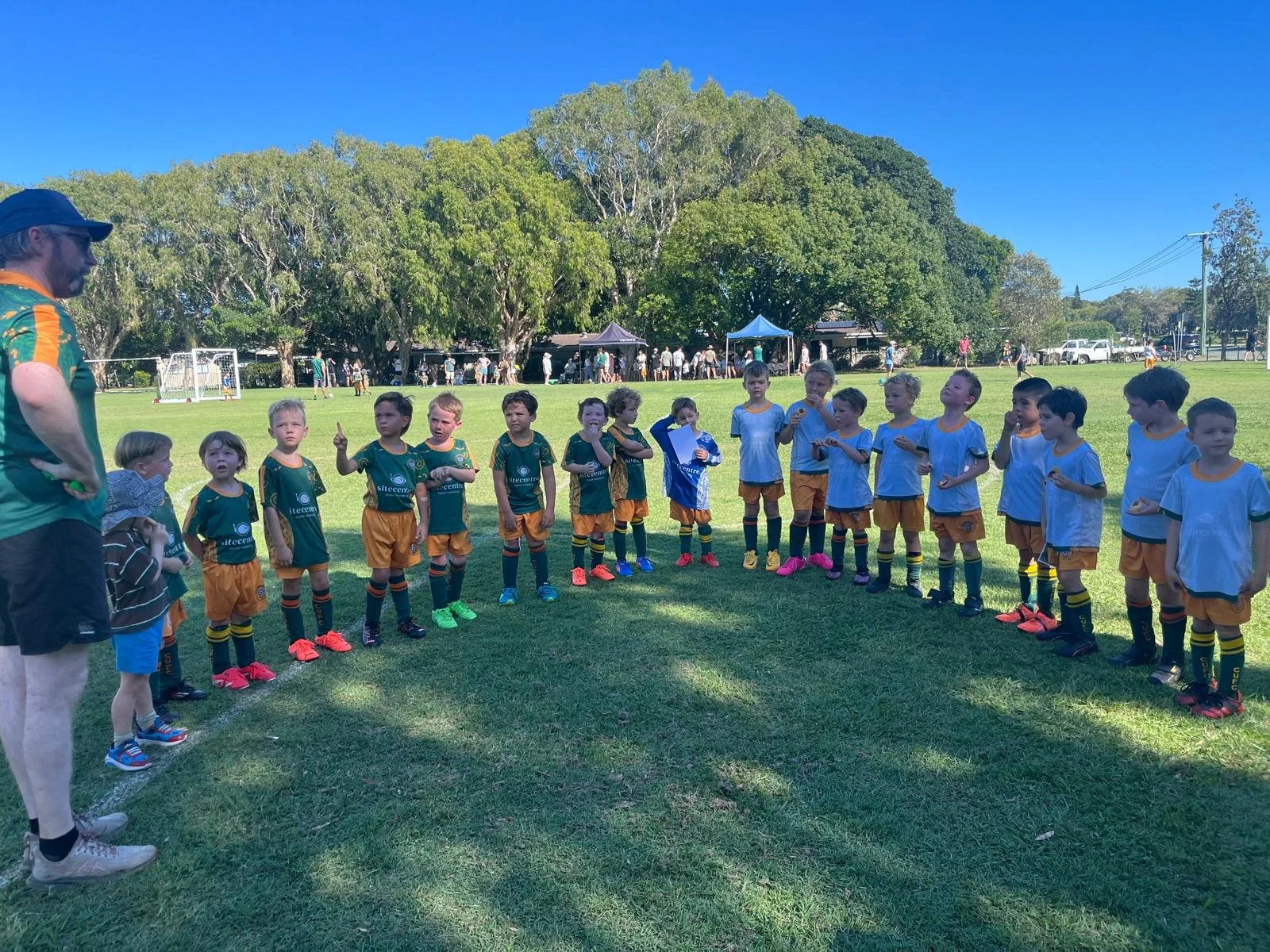 Children in sports uniforms standing in a line on a grass field, being instructed by a coach, with trees and tents in the background on a sunny day.