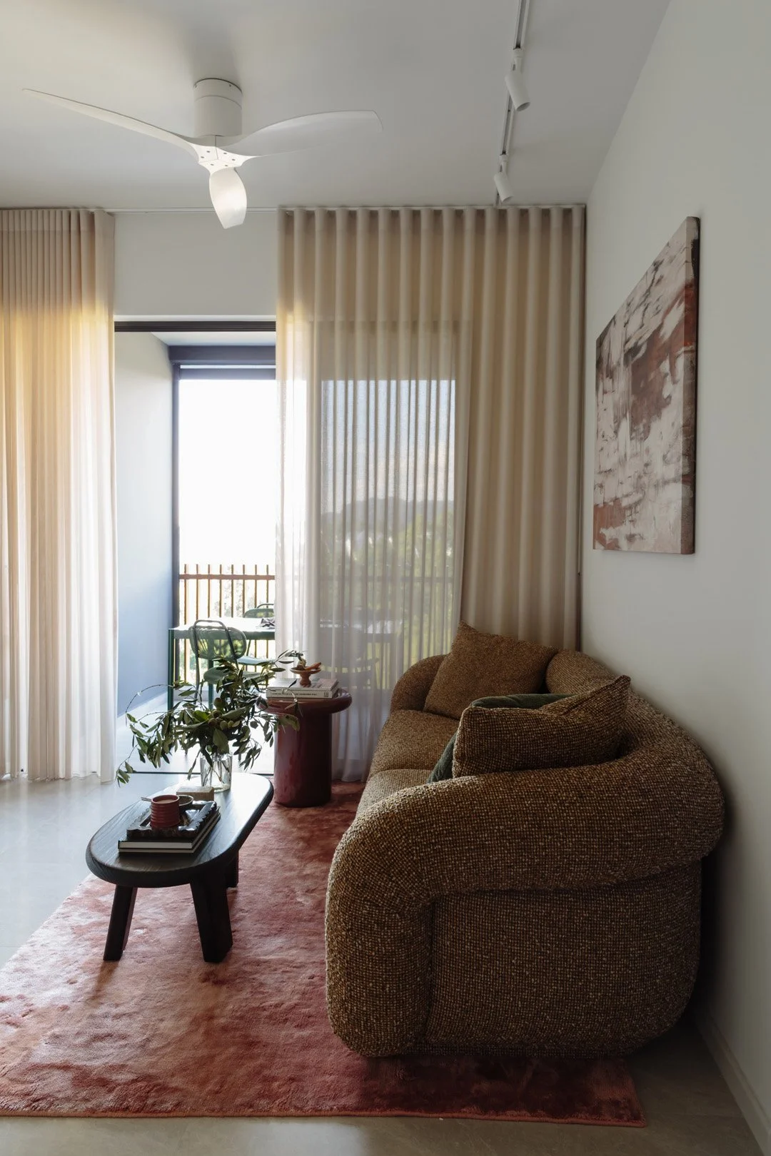Living room with a brown textured sofa, a black coffee table with a plant and books, a pink rug, and sheer curtains over a sliding glass door that leads to a balcony with green outdoor furniture.