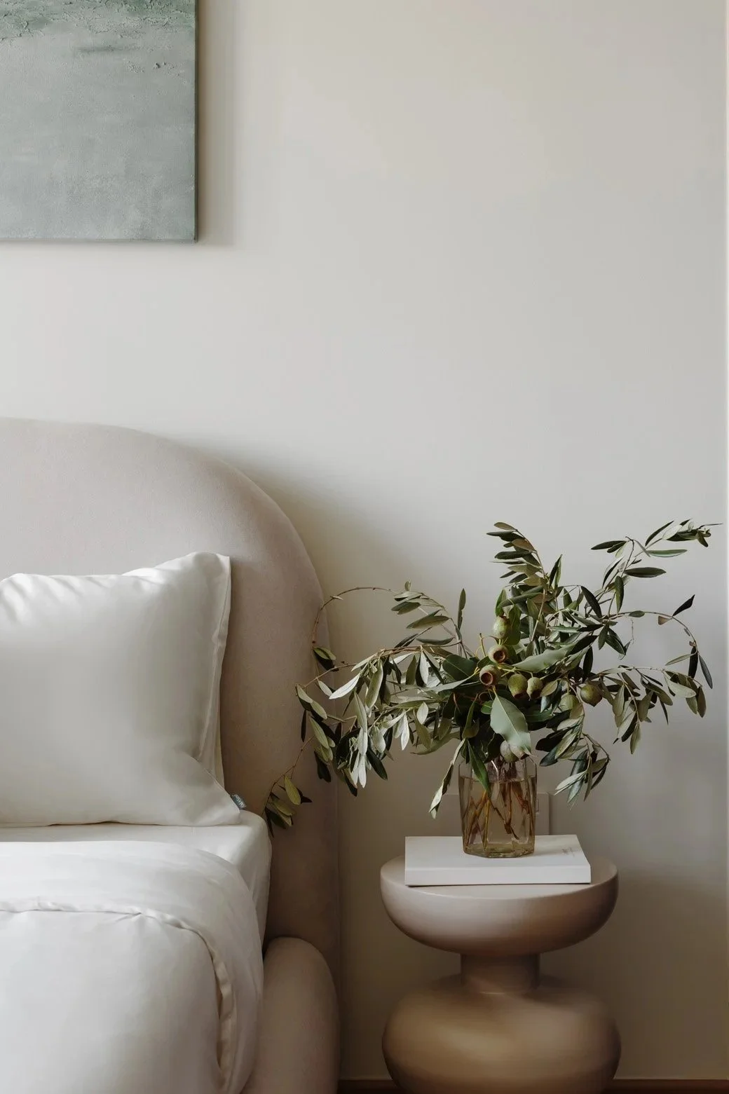 Living room corner with beige upholstered sofa, white pillow, side table with a glass vase of green leafy branches, and a white book, against a plain white wall with a corner of an abstract painting visible.