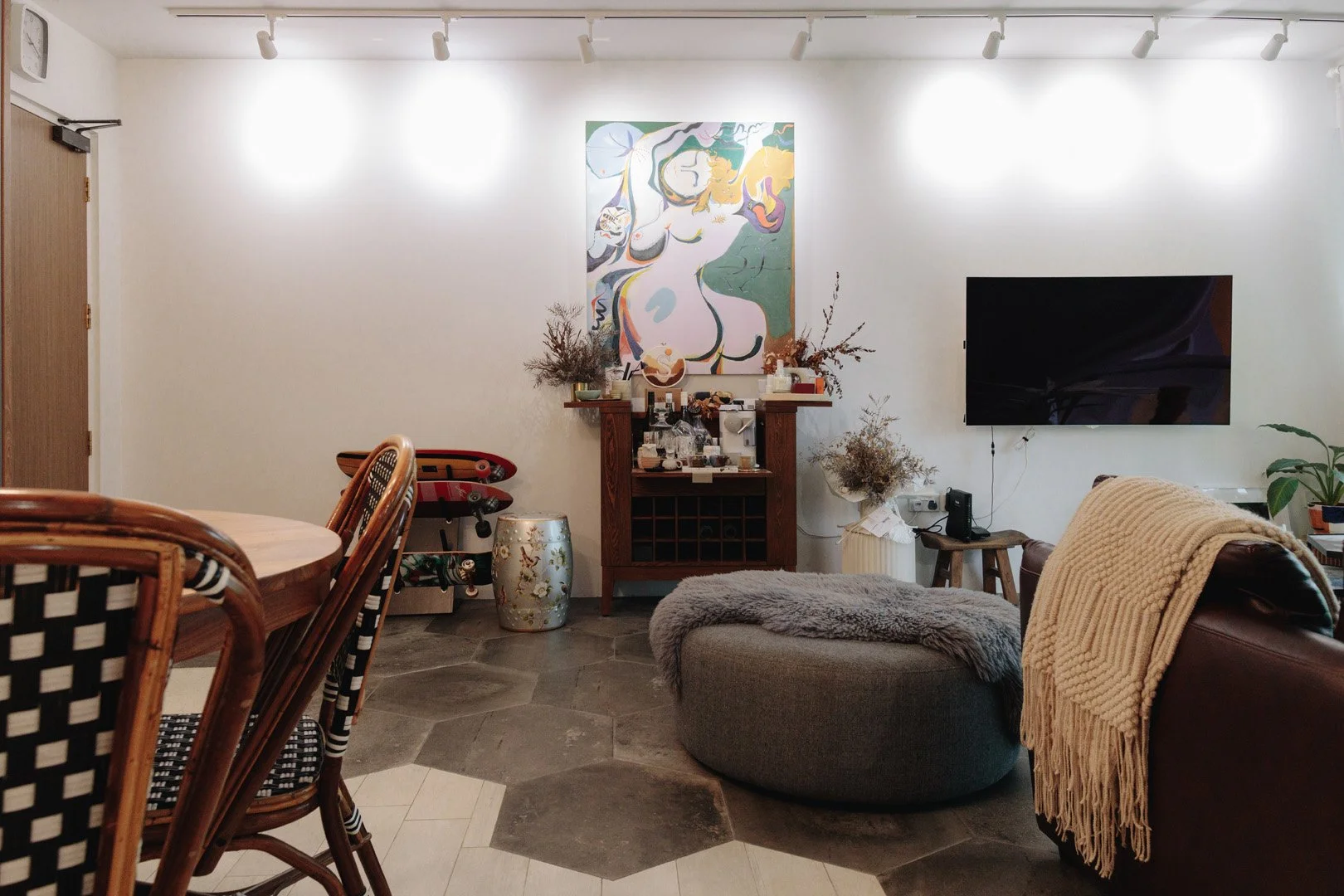 Living room with white wall, abstract artwork of a nude figure, a flat-screen TV, brown leather sofa with a beige blanket, decorative vase, plants, and a gray round ottoman on hexagonal tile and wood flooring.