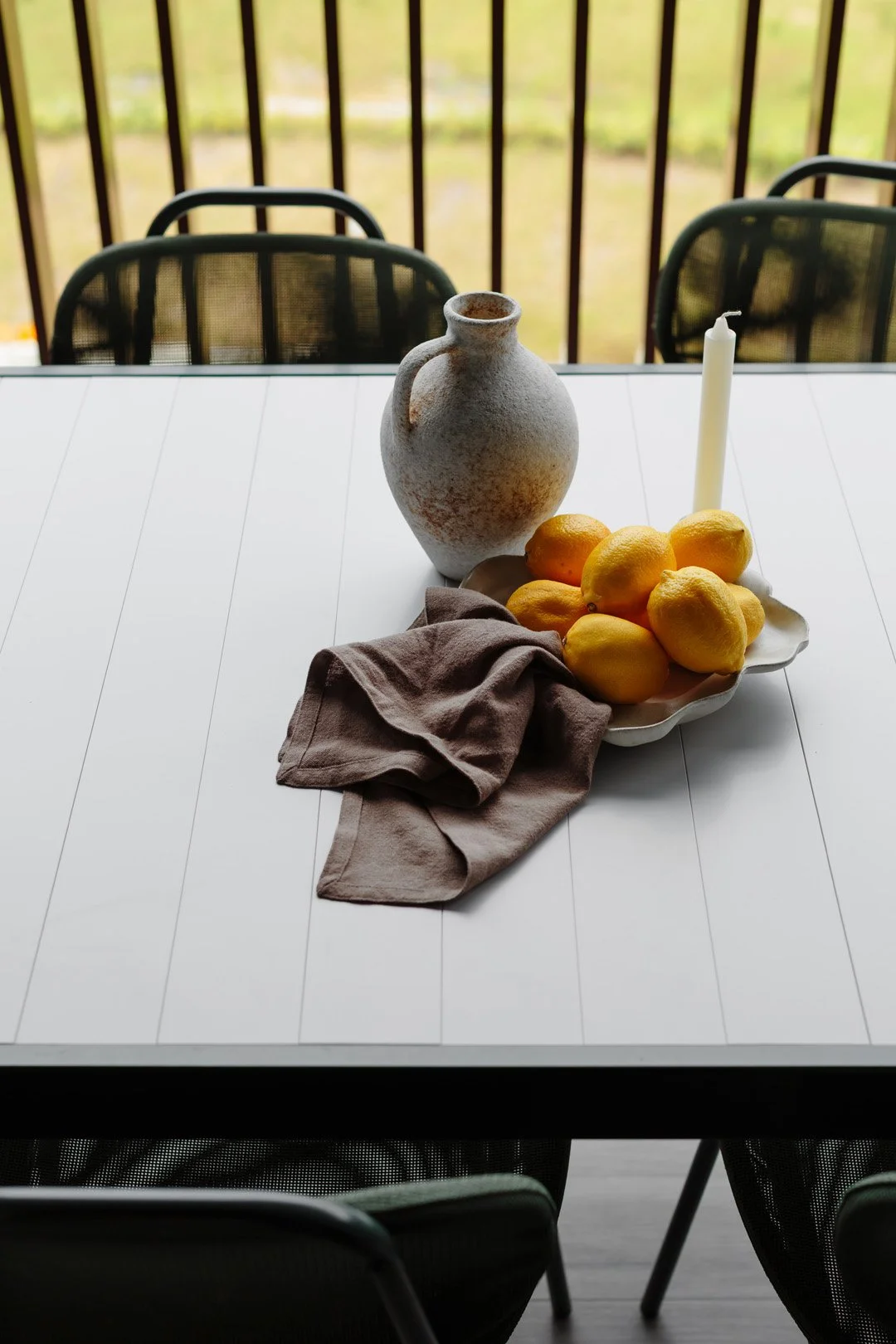 A white dining table with a ceramic vase, a plate of yellow lemons, a beige cloth, and a white candle, with a balcony and outdoor greenery in the background.