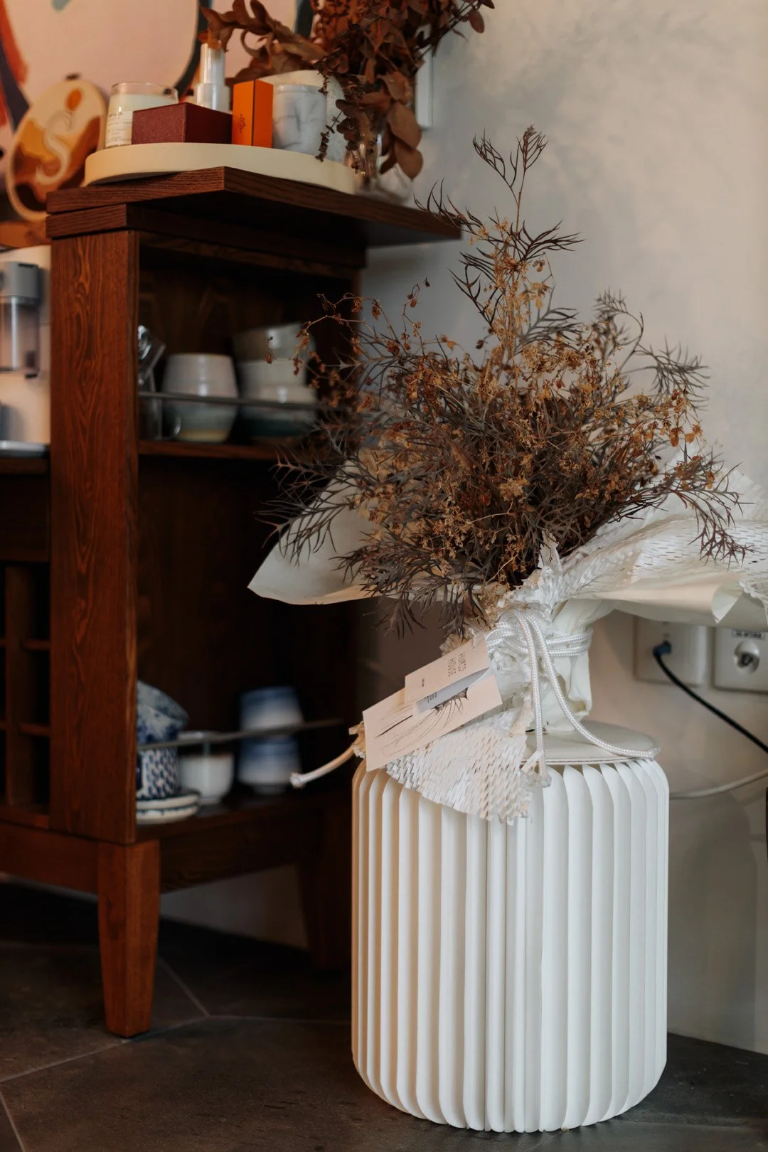 A white, ribbed ceramic vase with a bouquet of dried brown foliage, wrapped in white paper with tags, placed on a dark tile floor next to a wooden cabinet with dishes and cups.