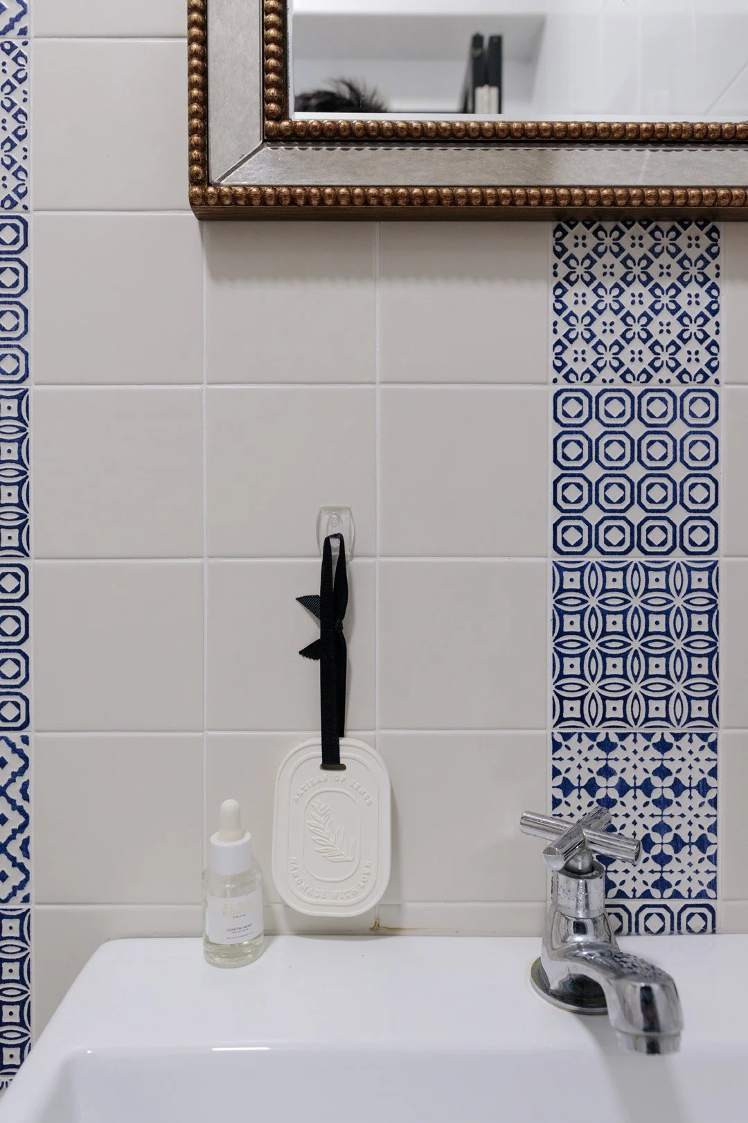 Bathroom wall with white tiles and blue patterned accent tiles, a mirror with a beaded copper frame, a soap bottle, a rectangular soap holder with a black ribbon, and a silver faucet.