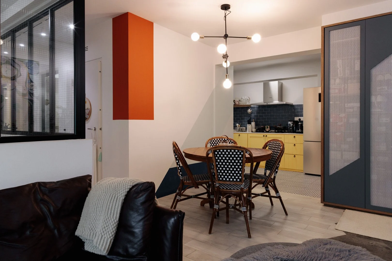Open-concept kitchen and dining area with a round wooden table, six black and white patterned chairs, yellow kitchen cabinets, black subway tile backsplash, stainless steel appliances, and a modern light fixture with exposed bulbs.