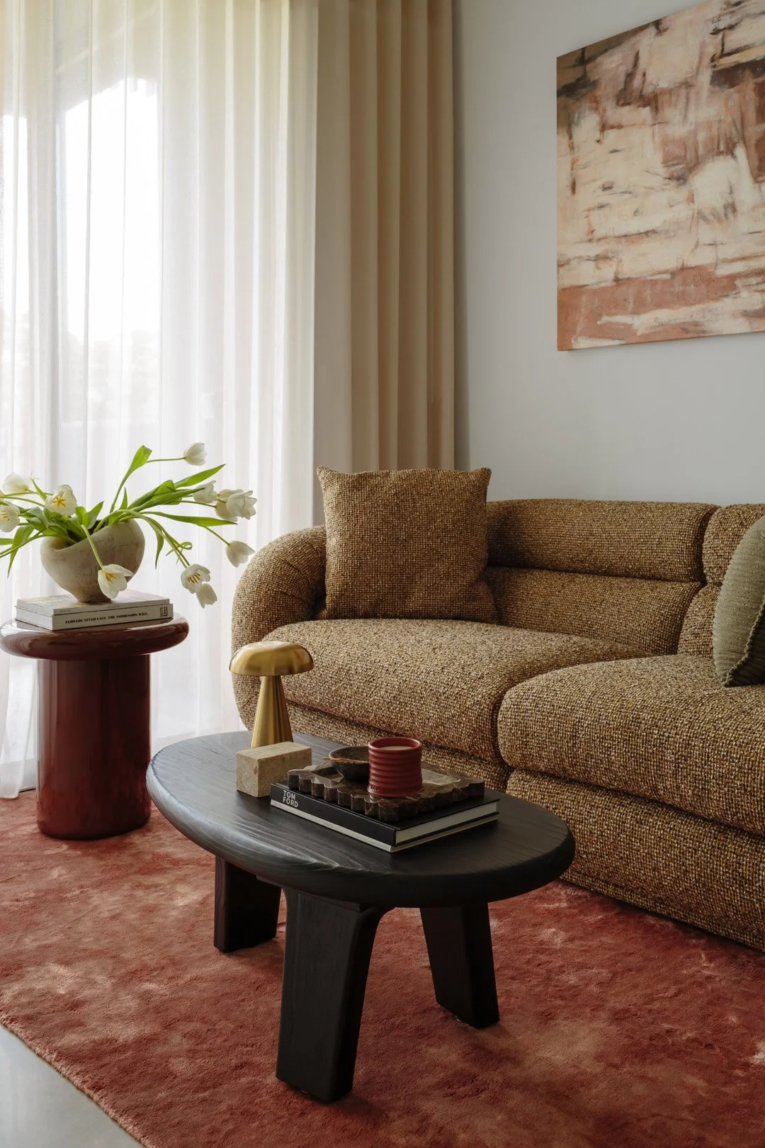 Living room with beige sofa, black coffee table, pink area rug, side table with white tulip flowers and books, abstract wall art, and beige curtain.