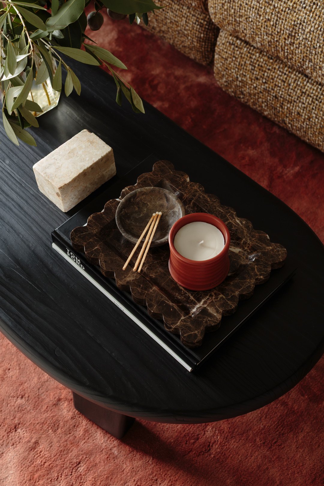 Decorative table setting with a candle in a red holder, an empty glass dish with chopsticks, on a marble tray, placed on a black wooden surface. A stone block and green plant are nearby, with a textured couch and red carpet in the background.