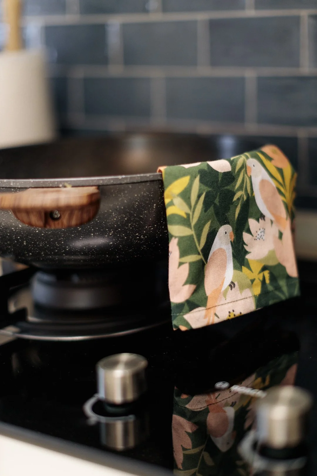 Close-up of a speckled black frying pan on a stovetop with a colorful kitchen towel featuring parrots and green leaves hanging over the side of the pan.