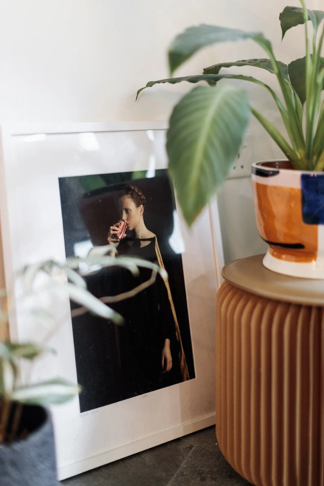 A framed photograph of a woman with braided hair drinking from a can, placed on the floor next to a round ribbed wooden side table with a colorful ceramic pot featuring a green plant with large leaves.