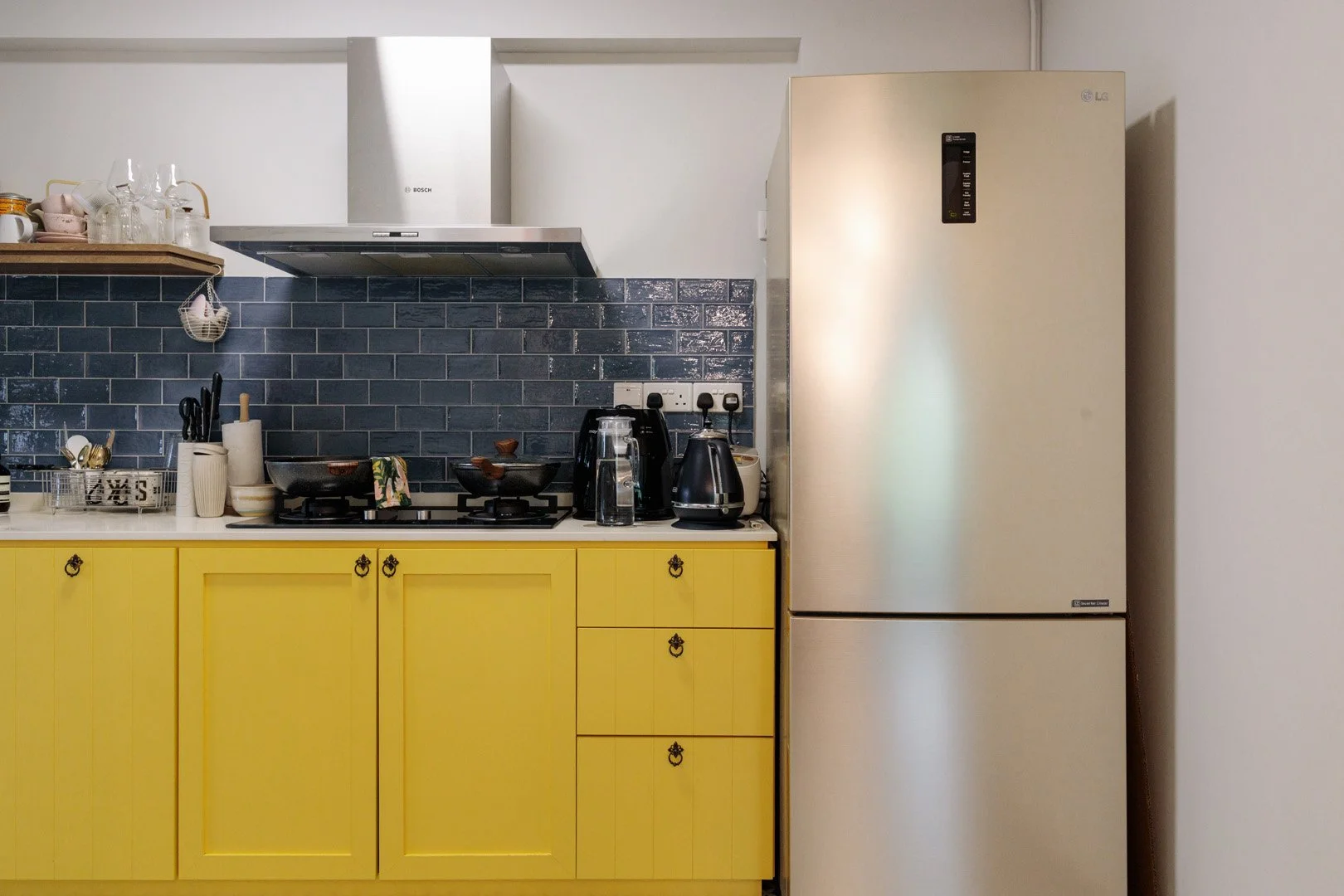 Kitchen with yellow cabinets, a black countertop with two stove burners, black kitchen appliances, a stainless steel refrigerator, a black tiled backsplash, and various kitchen items on the countertop.