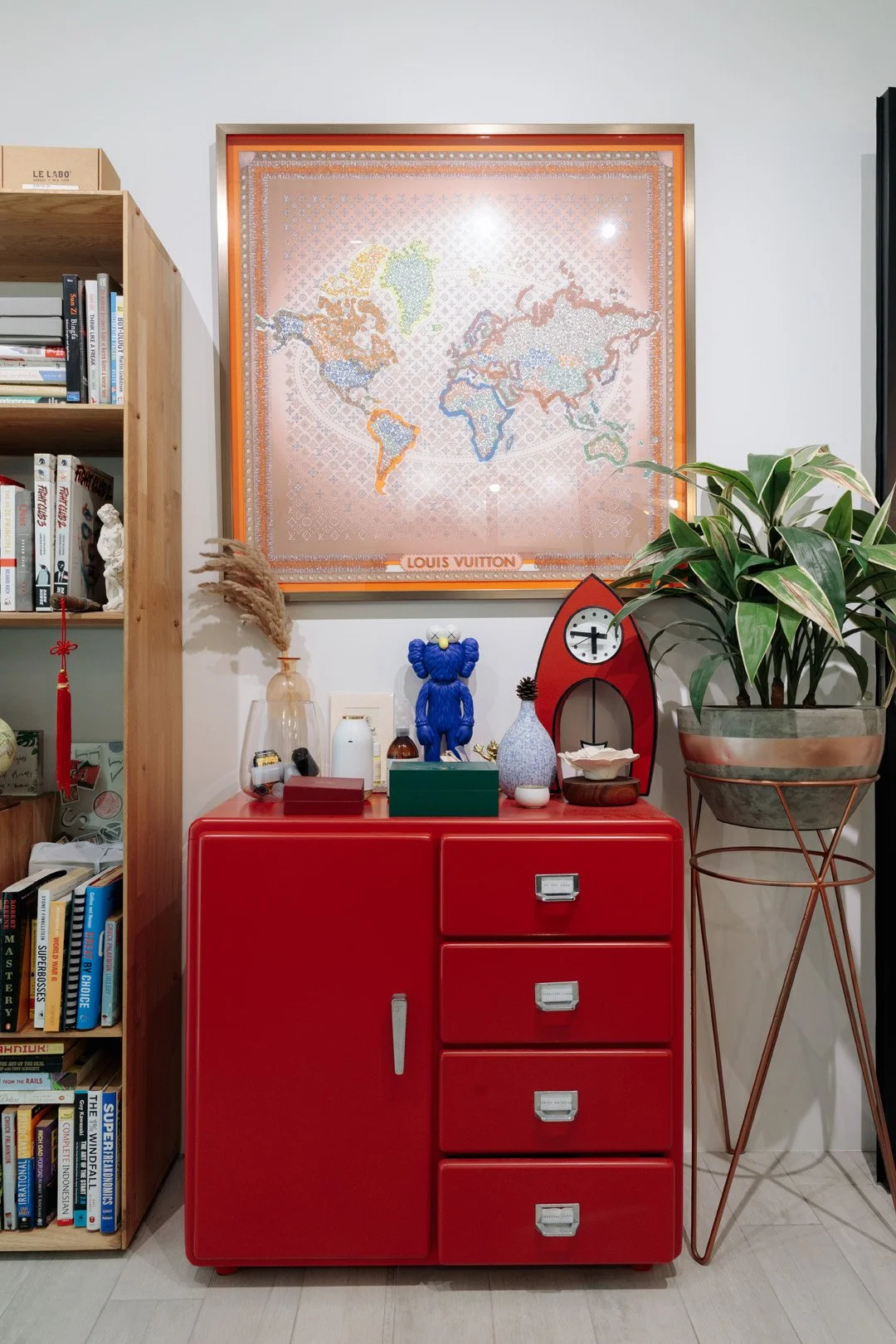 A red cabinet with four drawers and a door, topped with various decorative items, beside a large plant in a pot, against a white wall with a framed world map and a bookshelf on the left.